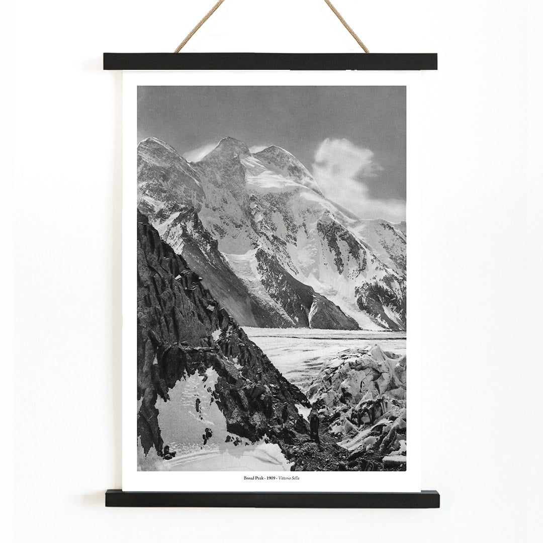 Black and white photograph and poster of Broad Peak by Vittorio Sella showing snow covered mountains and mountaineers climbing amidst rocky terrain and glacier, with wooden magnetic frame on white background